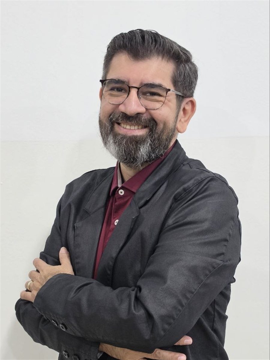 Josué Ribeiro sorrindo, de braços cruzados, vestindo blazer escuro e camisa vinho